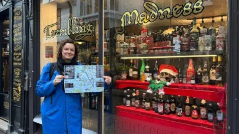 University of York Victoria Wells smiling and holding up the map, wearing a long blue coat and stood outside the House of Trembling Madness bar, where there are several bottles of alcohol in the window as well as a skull with a Santa hat on. 