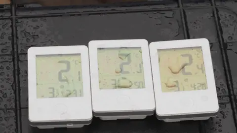 BBC/Mark Ansell The photograph shows three small digital thermometers placed side by side on a dark, wet surface. All three thermometers are rectangular with white casings and digital displays.
Each display shows temperature readings in degrees Celsius: 2.1, 2.1, 2.4.