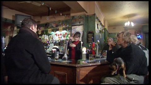 Barmaid stood behind a bar pulling a pint.  Punters in the foreground