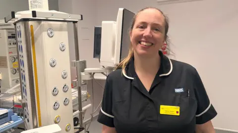 Head of midwifery Kerry Taylor smiling for the camera. She is wearing a black nurse's uniform. She has long fair hair tied back in a ponytail. Next to her are some pieces of medical monitoring equipment.