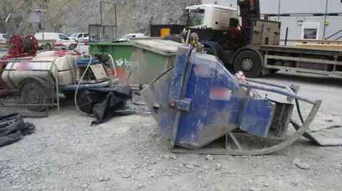 HSE A large blue metal skip is lying on the ground on its side. There are various pieces of industrial equipment around it. A lorry is parked nearby.