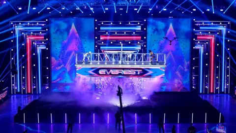 BBC/Hungry Bear Large illuminated stage with vibrant neon lights, digital mountain graphics, fog effects, and a raised platform displaying the word ‘Everest,’ with several figures standing on the platform during a live show or performance