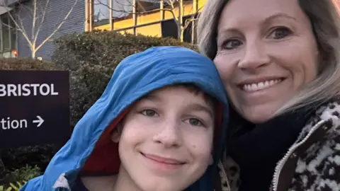 Katharine Theo on the left and his mum Katharine on the right. Both are smiling at the camera. Theo has the hood of a blue coat over his head. There is a large building and shrubbery in the background.