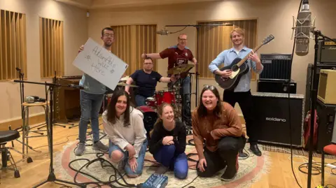 Band of seven people posing in front of a drum kit and speakers while smiling. There are in a room with blinds on the windows. The person on the left is holding a whiteboard which reads: "AutismAble was Here". The person on the right is holding a black guitar. There are three women crouched in front of the drum kit. The man at the drum kit is smiling in a black top and holding the cymbal. There are wires strewn across the floor.