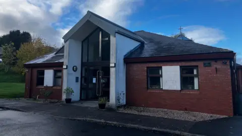 A brick building with a white opening in the front. the top of the door is adorned with a big silver cross.