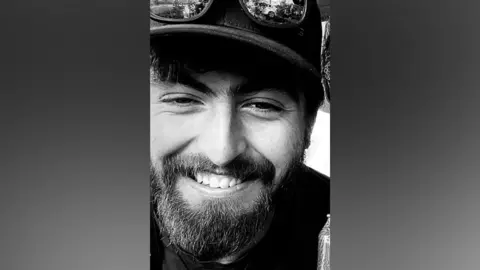 Byrne family A black and white image of smiling Liam Byrne who has a dark beard and is wearing a black cap