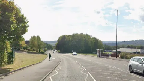 A wide single carriage road with just two or three cars on it bends between trees into the distance. A single pedestrian walks on one of the wide pavements on either side of the road.
