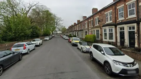 A Google Street View screenshot looking up Greenbank Road in Darlington. There are terraced houses with bay windows on the right and a green space with trees on the left. Cars are parked at either side of the road.