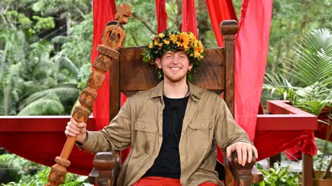 James Gourley/ITV/Shutterstock AngryGinge grinning on his throne, with a sunflower crown on his head. He is also holding a wooden staff.