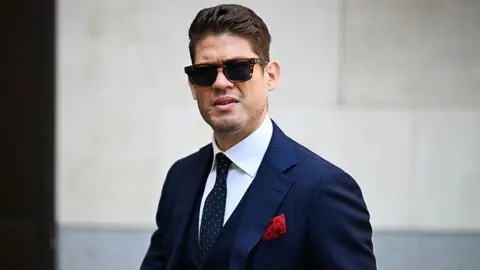 A man with short brown hair wears dark sunglasses and a blue pinstriped suit with a red pocket square looks without smiling towards the camera.