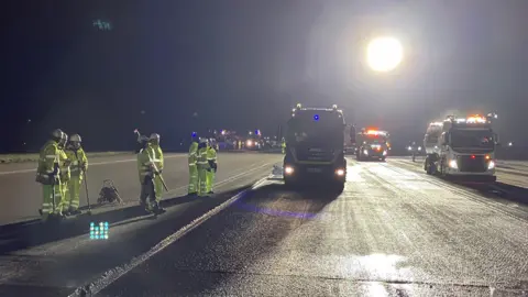 BBC/SAM READ The image shows a well-lit airport runway at night with a large floodlight at the back of the picture. There are three large construction vehicles in the foreground with a group of workers in high-visibility clothing standing to the left side of the picture. There are a series of small lights in the distance. 