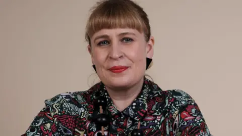 Polly Garnett A woman with blonde hair tied back. She is wearing a multi-coloured patterned top.