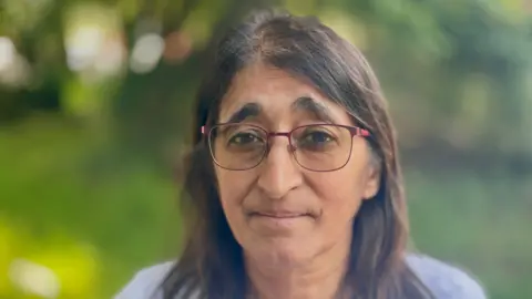 Stephen Huntley/BBC A woman with long dark hair and glasses and wearing a grey top smiles at the camera. She is smiling. She is outside. The background is blurred.