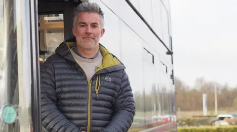 LDRS A man looking straight ahead. He is stood in the doorway of a bus and is wearing a black padded jacket with green lining. 