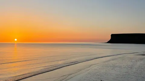 Sarah n Essie An orange sunset at the beach in Saltburn with the sun reflecting beams off the waves. The silhouette of a nearby cliff can be seen on the right of the picture. The sun is about to set on the left.