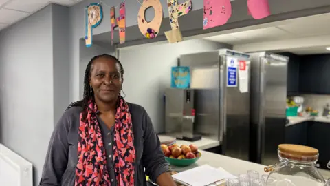 Jon Wright/BBC Mollin Delve stands by a kitchen counter hatch. There are fridges in the background and a bowl of apples.