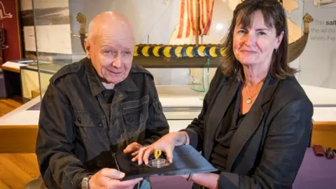 Manx National Heritage/Simon Park Ronald Clucas and Allison Fox stand holding the the arm ring, which is a small piece of gold on a stand. Allison wears a dark blazer and top, with a silver necklace. She has shoulder length brown hair. Ronald is bald and wears a dark jacket. They are in front a museum display of a Viking longboat.