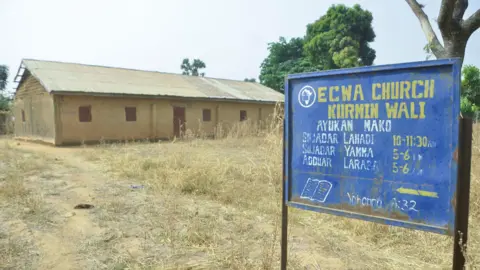 Exterior view of ECWA Church, after an attack by gunmen in which worshippers were kidnapped. The church is made of brown bricks. There is a blue sign with the name of the church and service times in yellow. 