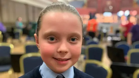 A girl with light brown hair tied back from her face is looking at the camera. She is wearing a navy v-necked jumper over a pale blue shirt with a navy and pale blue striped tie