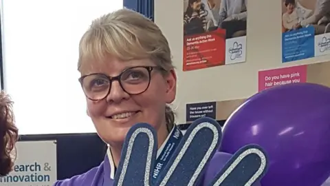 East Kent Hospitals A blonde woman wearing glasses in purple nurses scrubs holds a foam hand and balloon. She is smiling at the camera. 