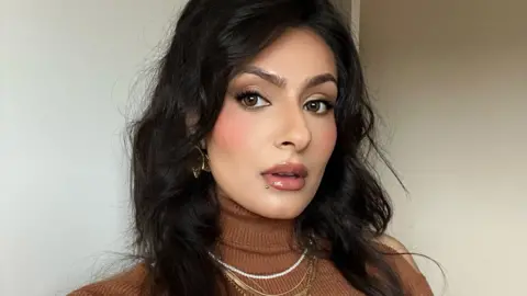 Iqra Qadir A woman wearing a brown roll neck jumper and gold jewellery looks into the camera.