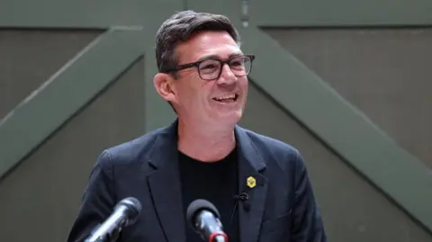 Andy Burnham is standing in front of a green background. He is a white man with dark hair and is wearing glasses, a black t-shirt and blue jacket with a yellow bee pin badge.