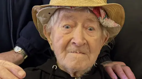 Ron "Ginger" Parsons looking at the camera, he is wearing a straw hat and a black hoodie. He has white hair. 