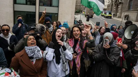 A group of protesters stand on a street corner in London. Some of them are shouting and smiling with their hands in the air. Many are holding phones up, one is holding a camera with a fluffy microphone and one has a megaphone.