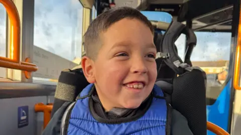 A young boy sitting on a bus and smiling. He has short dark brown hair and brown eyes and is wearing a padded blue vest and a black jacket.