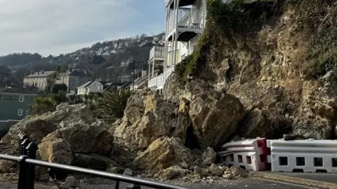 Rocks and debris strewn across Belgrave Road, and at the back of the road, plastic bollards have been crushed by the weight of rocks on top of them. Buildings built alongside the cliff face are visible further away. 