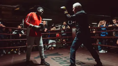 Paul Gilfeather Frank en un ring de boxeo con un chándal oscuro y también lleva guantes de boxeo marrones con coronas doradas. Frente a él está uno de sus estudiantes vestido con pantalones deportivos grises y una blusa roja. Parece estar mostrándole técnicas de boxeo y detrás de las cuerdas del ring hay estudiantes observando la demostración.
