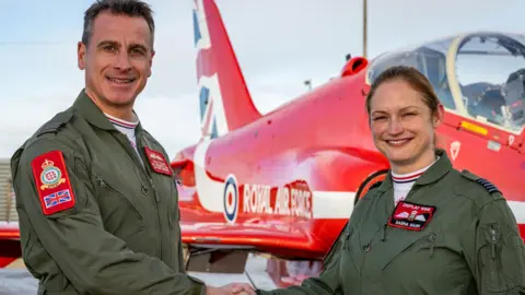 MOD Wing Cdr Adam Collins and Wing Cdr Sasha Nash standing in front of a Red Arrows jet. They are dressed in flying overalls and are shaking hands while smiling at the camera. 