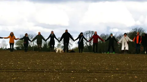 Block East Pye Solar About 10 people hold hands in a ploughed field. A dog is standing in front of them.