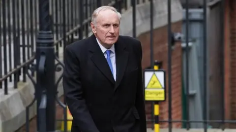 PA Media Dacre walking in front of a black metal fence. He wears a long black coat, a white shirt and blue tie. He looks sincere 