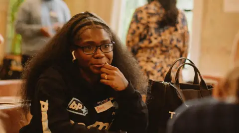 Hannah Henderson, Settle Jade Barnett has long brown hair and is wearing glasses and a black jukped with various logos on it. She is sitting at a table with one hand close to her face. There are a couple of people in the background.