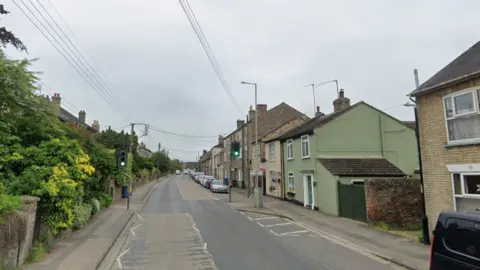 Google A view of Melford Road in Sudbury. Houses line the street on each side. A pedestrian crossing with traffic lights can be seen. 