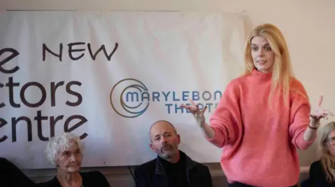 Gavin Watson Woman in pink jumper with blond hair addresses audience in front of a sign for The New Actors Centre at Marylebone Theatre
