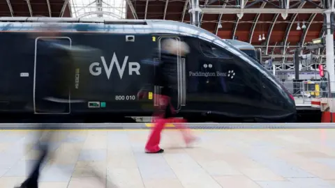 REUTERS/ Belinda Jiao A dark green Great Western Railway train sitting in London Paddington. The train is called Paddington Bear and there is a pedestrian wearing red trousers walking in front of the train. 