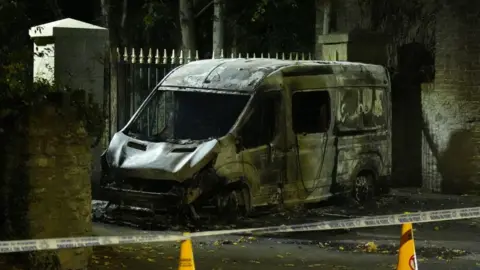 PA Media The wreckage of a burned out garda van in Dublin. It is windowless and has scorch marks. There is police tape nearby and yellow cones.
