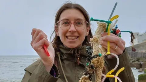 A woman with brown hair and glasses smiling at the camera. She is wearing a green coat and is holding sea plastic and waste up either side of her face. The sea is behind her