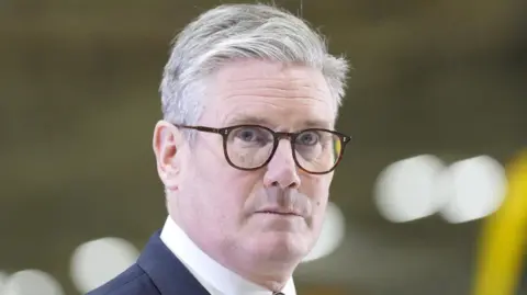 Keir Starmer, with grey hair, looking to the side of camera. He is wearing brown glasses.