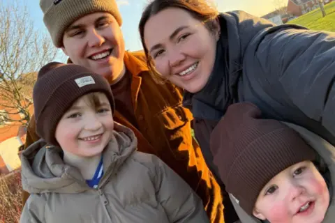 Ella Ella Nickson and her partner Jae withe their two children are dressing in warm coats and warm hats and stood in a group as they smile at the camera outside