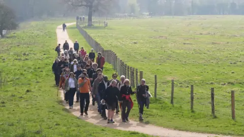 PA Media Roughly 30 people walking towards the camera, along a mud path through a grassy field.