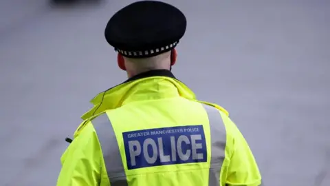 A police officer facing away from the camera wears a police hat and a yellow jacket with the words 'Greater Manchester Police' on the back 