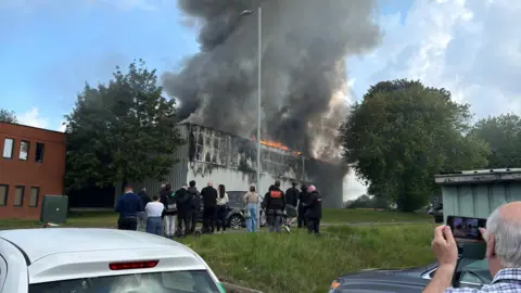 A crowd of people gather on a grassy bank as they watch a grey building with orange flames on the roof. A large plume of dark smoke is rising into the blue sky. 