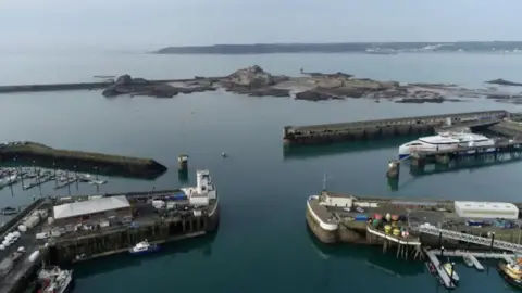 BBC An aerial view of the Ports of Jersey - four separate walls sticking out in the water with boats surrounding, in the background you can see Elizabeth Castle and further afield is the coastline of Jersey.
