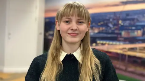 BBC A female pinch blonde hairsbreadth smiles into nan camera. She is wearing a bluish and greenish coloured jumper pinch a garment underneath. She is opinionated successful agency successful beforehand of an image depicting a metropolis skyline.