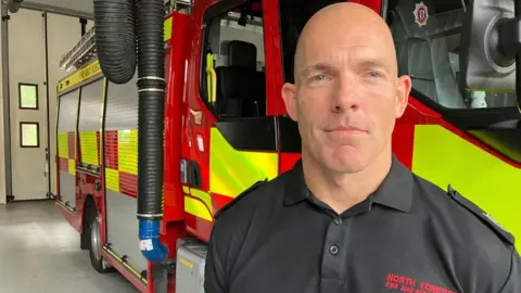 A man wearing a fire service polo shirt looks into the camera with a neutral expression on his face. He is standing in a large garage with a fire engine behind him.