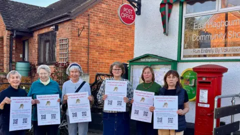 A group of six female campaigners holding up placards in front of East Hagbourne Community Shop - which is a white building with a post box outside.
