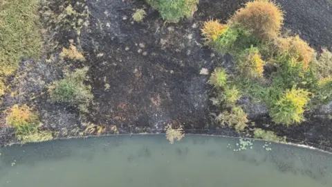 Northamptonshire Fire and Rescue Service A scene from above of a burnt field with trees all around it, and a large area of water. You can see burnt crops and murky water. 